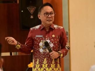 Seorang pejabat pemerintah daerah mengenakan batik marun sedang memaparkan materi di depan peserta forum.