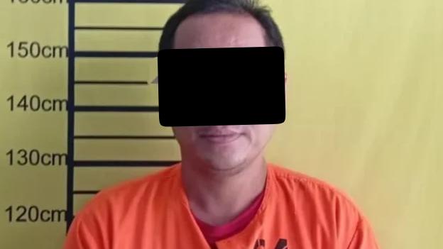 Tersangka kasus narkotika berompi tahanan berdiri di depan papan pengukuran tinggi.